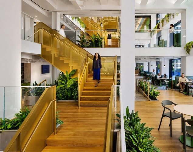Biophilic workplace design featuring integrated interior landscaping surrounding a brass staircase within a Manhattan corporate headquarters.